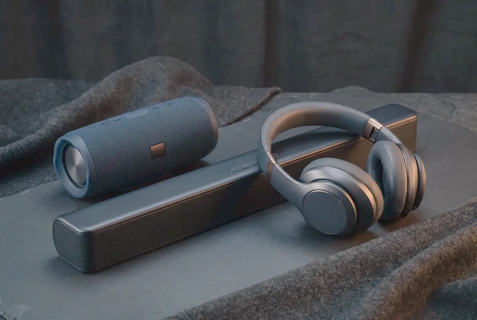 A portable speaker, soundbar and over-ear headphones on dark slate