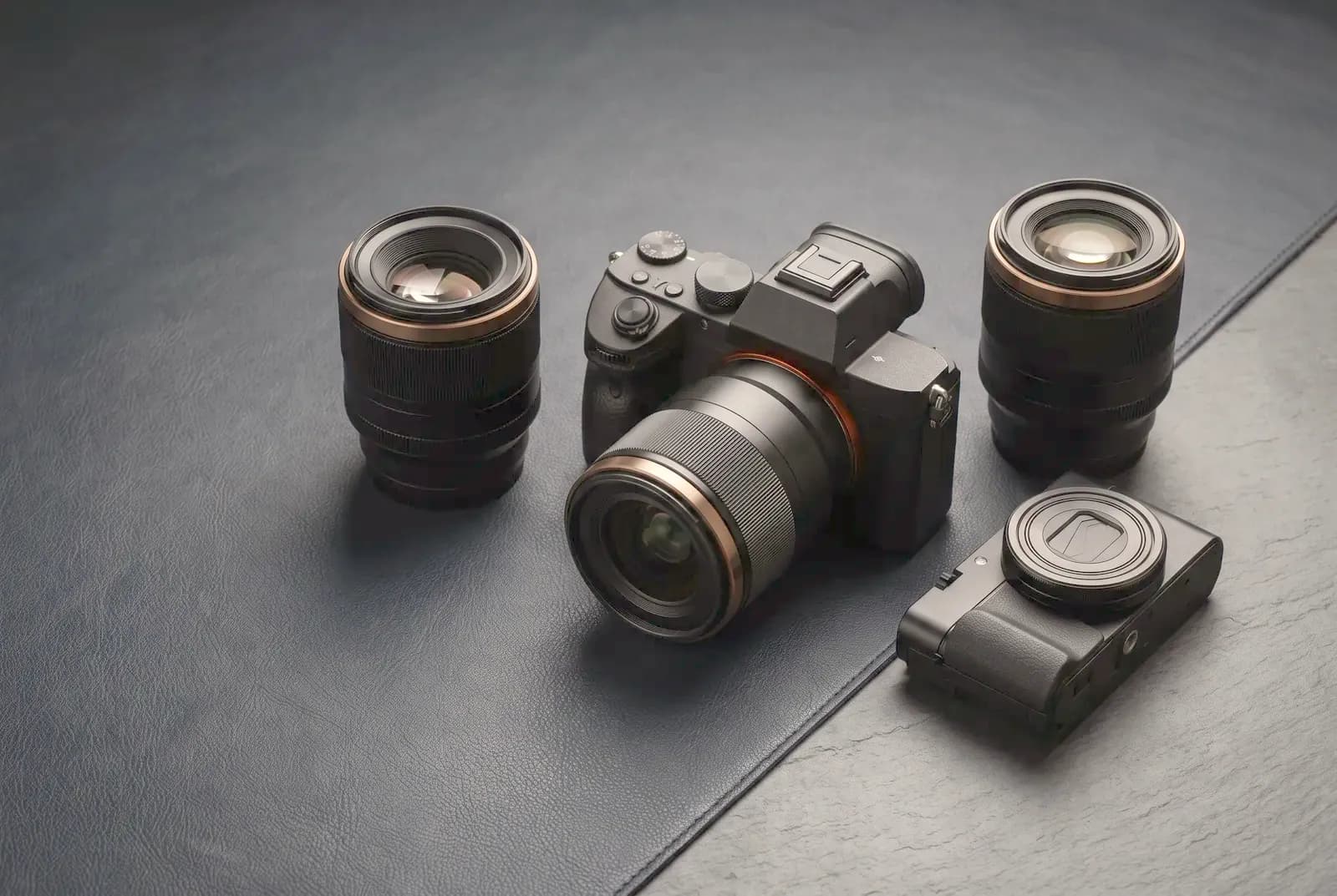 A mirrorless camera with lenses and a compact camera on leather and slate