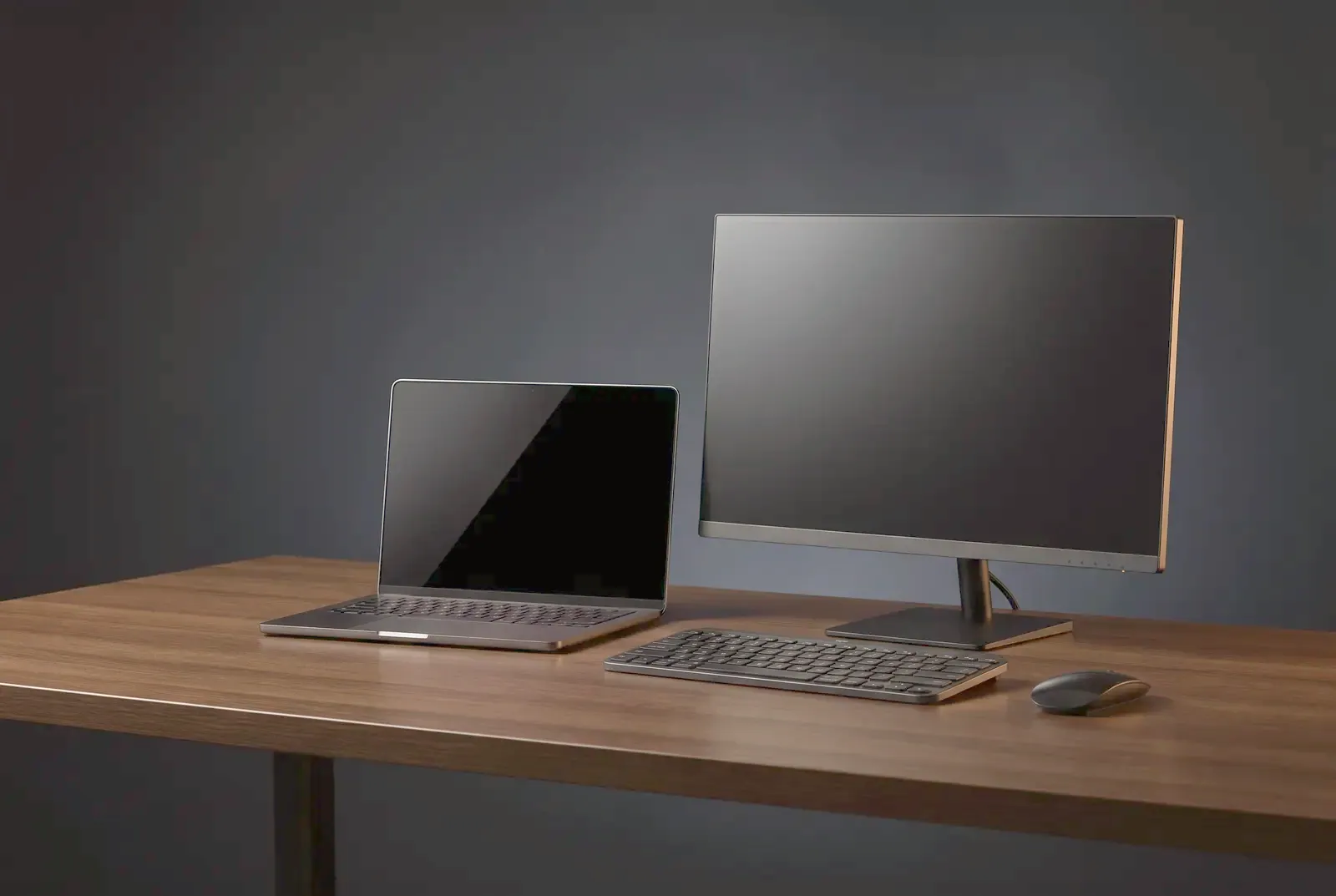 A slim laptop, monitor, keyboard and mouse on a dark oak desk